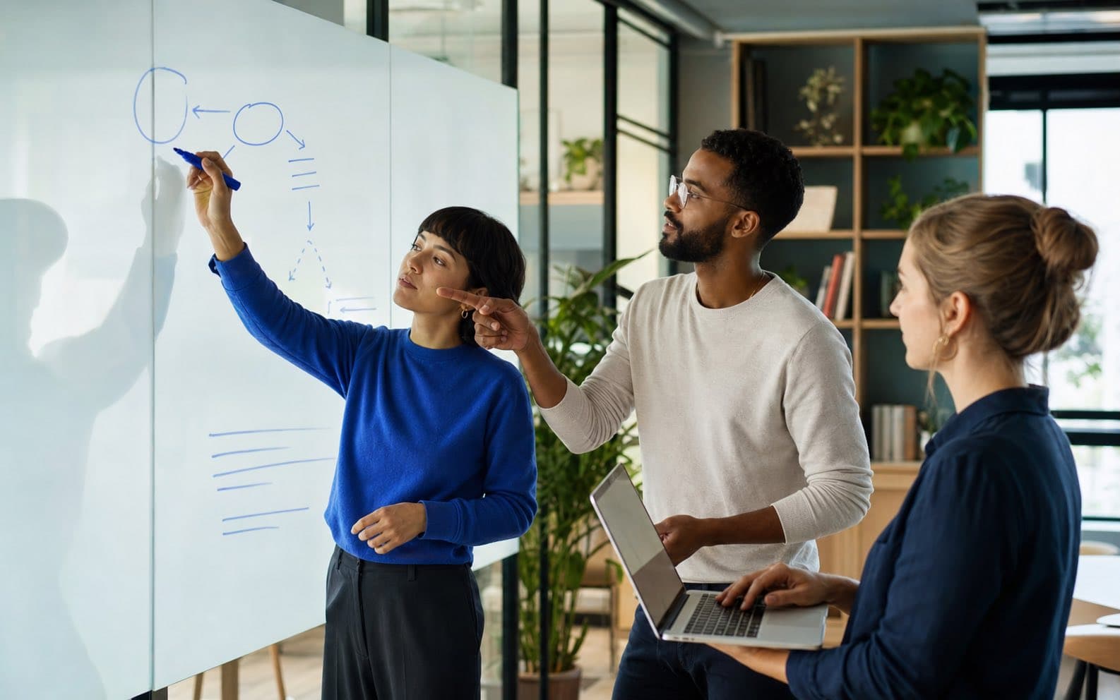 Three coworkers preserve the reasoning behind a decision using a diagram and a laptop in an office meeting room.